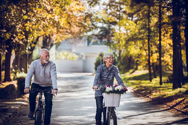 A retired couple riding bicycles down a quiet, tree-lined street in the fall.